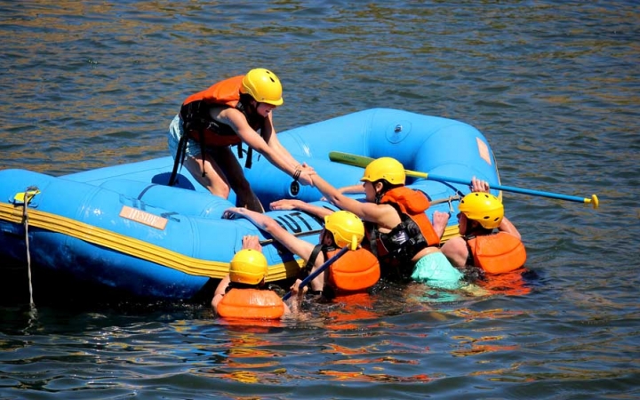 One person in a raft helps others who are in the water into the raft. All are wearing helmets and life jackets.